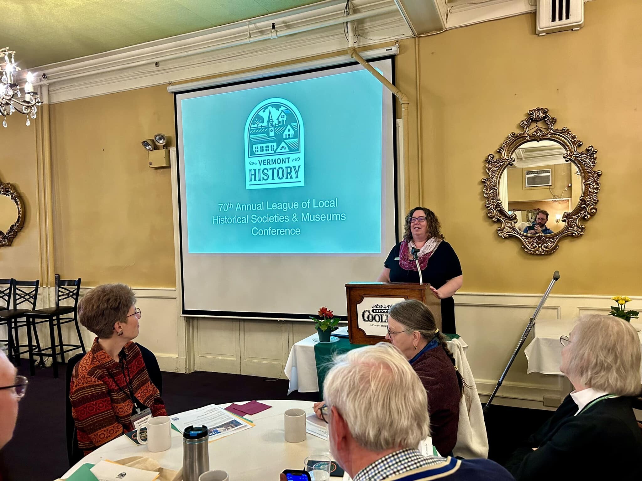 A person standing at a podium in a crowded room. The screen in the back reads 70th annual League of Local Historical Societies and Museums Conference