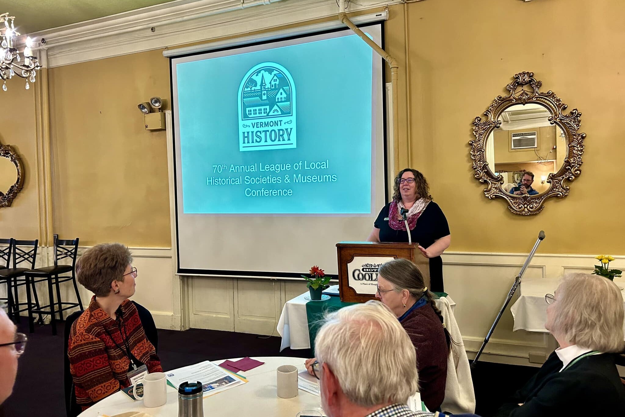 A person standing at a podium in a crowded room. The screen in the back reads 70th annual League of Local Historical Societies and Museums Conference